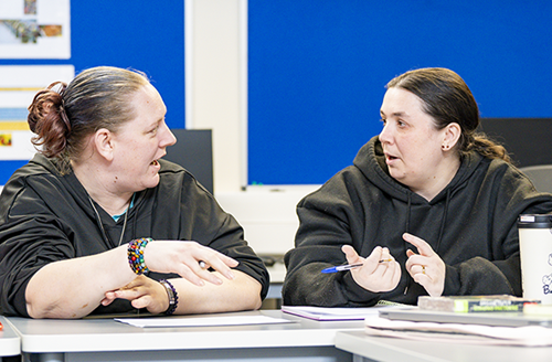 Kalie, with her classmate, Elizabeth in their class. They are sat at a table, with books in front of them. They both have their hair tied up, wearing black hoodie jumpers. They are engaging in a conversation.
