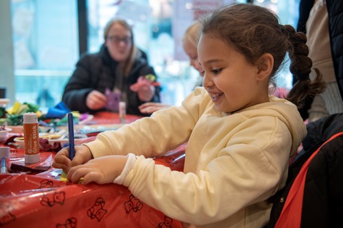 Young girl doing crafts, holding a blue pen. She is wearing a yellow jumper and has her hair in a brunette plait. In the background, a mum and her daughter are doing the same craft.