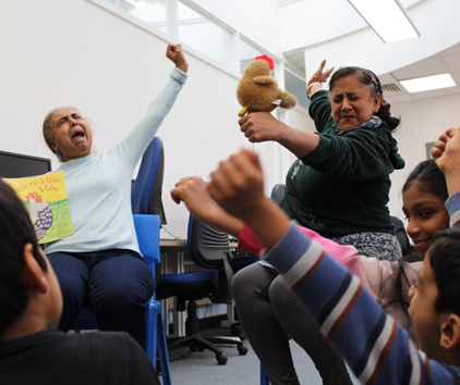Leicester Libraries Reading a book to children, acting out a hen 'waking up', with their arms up and yawning.